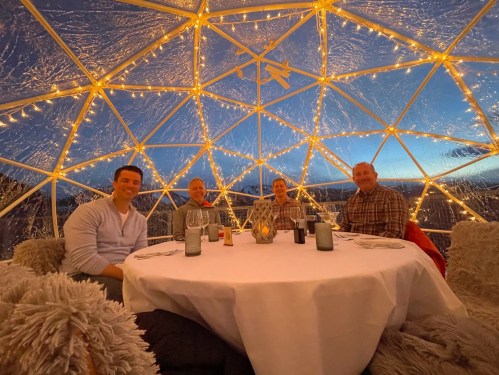 Four people sit around a table inside a cozy, illuminated dome, with a scenic view outside at dusk.