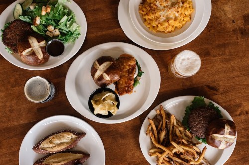 A table spread with salads, mac and cheese, burgers, fries, and drinks on white plates against a wooden background.
