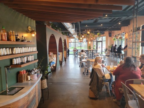 A cozy café interior with wooden accents, tables, and patrons enjoying drinks in a bright, inviting atmosphere.
