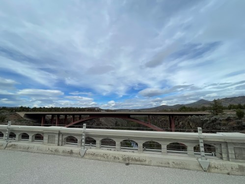 A wide view of a bridge spanning a valley under a cloudy sky, with mountains in the background.