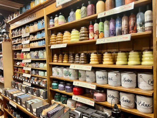 A display of various scented candles in jars and containers on wooden shelves, showcasing different colors and labels.