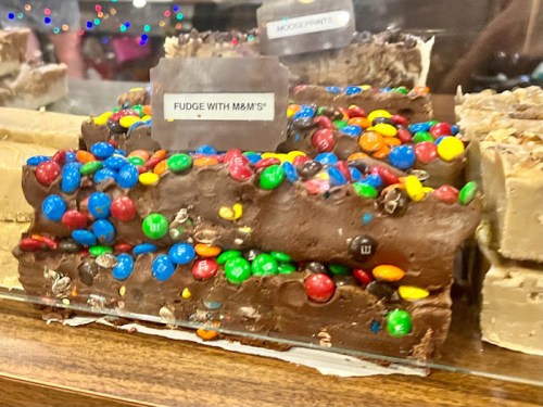 A close-up of fudge topped with colorful M&M's, displayed in a glass case with a label reading "Fudge with M&M's."