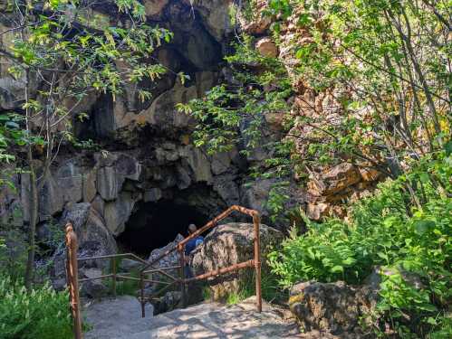 A person descends stone steps into a dark cave entrance surrounded by lush greenery and rocky terrain.