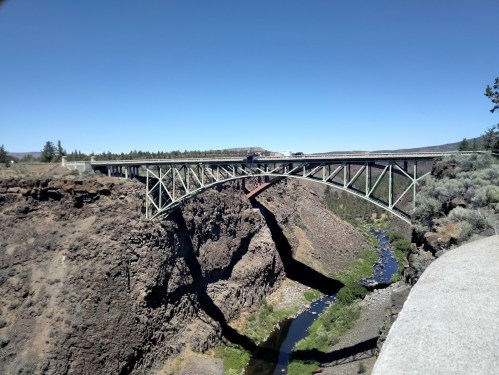 A green metal bridge spans a deep canyon with a river below, surrounded by trees and clear blue sky.