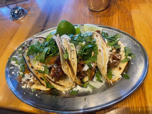 A platter of tacos topped with cilantro, onions, and cheese, served with lime wedges on a wooden table.