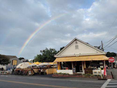 A charming storefront with a yellow-striped awning, bicycles outside, and a rainbow in the sky above.
