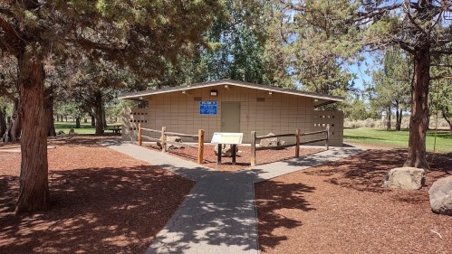 A small, light-colored building surrounded by trees and gravel paths in a park setting.