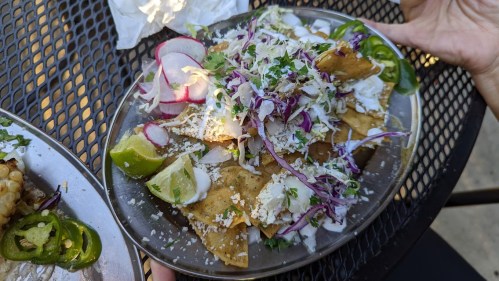 A plate of chilaquiles topped with radishes, cabbage, cheese, cilantro, and lime wedges, served on a metal tray.