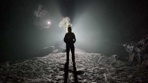 A silhouetted figure stands in a dimly lit cave, with beams of light illuminating the surrounding darkness.