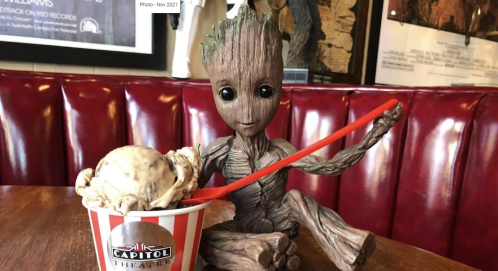 A wooden Groot figure sits at a table with a cup of ice cream, holding a red spoon, in a cozy diner setting.