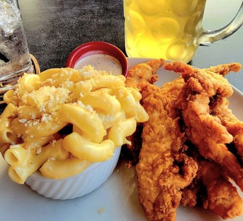 A plate with crispy fried chicken strips and a side of creamy macaroni and cheese, with a drink in the background.