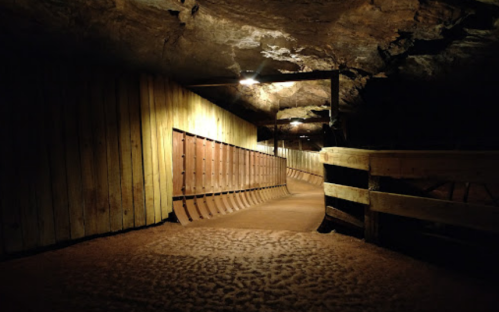 A dimly lit underground tunnel with wooden walls and a sandy floor, illuminated by overhead lights.