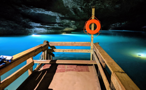 A wooden dock extends over a serene, illuminated blue underground lake in a cave. A life ring hangs nearby.