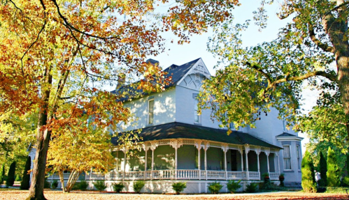A large, historic house surrounded by colorful autumn trees and a wraparound porch.