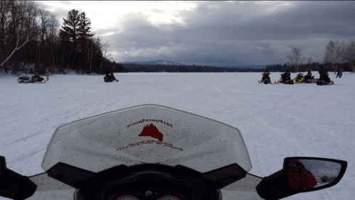 View from a snowmobile on a snowy lake, with other snowmobiles and trees in the background under a cloudy sky.