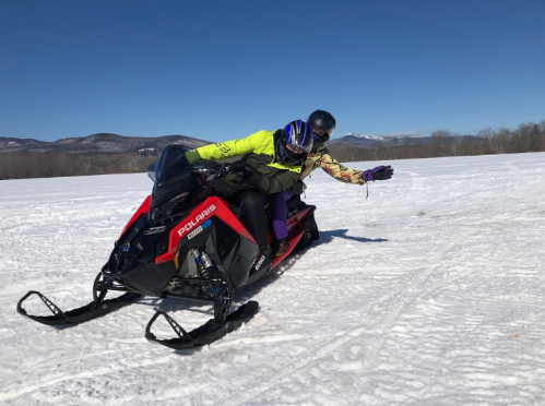 Two people riding a snowmobile on a snowy landscape with mountains in the background under a clear blue sky.