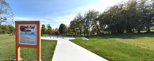 A splash pad area in a park, featuring a sign and water play equipment surrounded by green grass and trees.