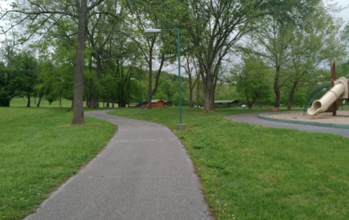 A winding path through a green park, with trees and a playground visible in the background.