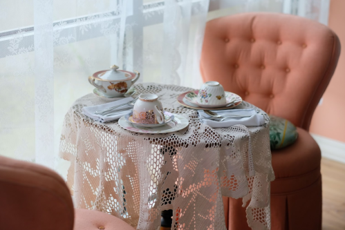 A cozy tea setting with delicate china on a lace tablecloth, surrounded by pink upholstered chairs by a window.