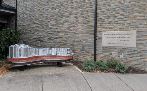 A sculptural bench resembling books sits outside a stone building, with a quote and date engraved on the wall nearby.