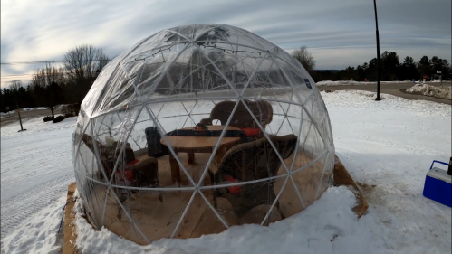 A transparent geodesic dome on snow, featuring a wooden table and chairs inside, set against a cloudy sky.