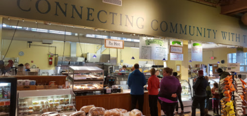 A bustling market with people in line at a food counter, surrounded by baked goods and a sign about community connection.