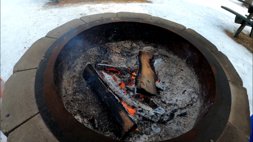 A fire pit with glowing embers and charred logs, surrounded by a stone border, on a snowy ground.