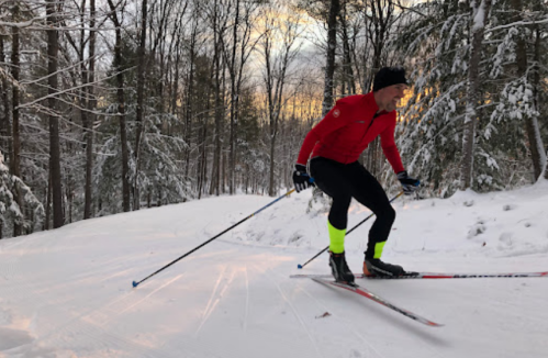 A skier in a red jacket and black pants glides through a snowy forest trail at sunset.