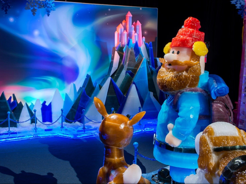 A colorful ice sculpture of a bearded man in a blue outfit and a red hat, with a deer and a mountain backdrop.
