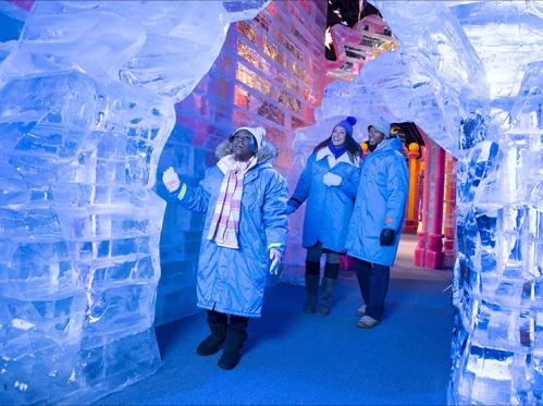 Three people in blue coats explore a colorful ice sculpture exhibit, surrounded by shimmering ice formations.