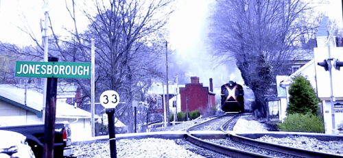 A train approaches a small town sign for Jonesborough, surrounded by trees and buildings on a cloudy day.