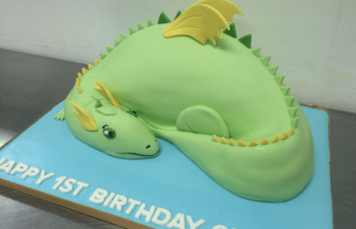 A whimsical green dragon cake, curled up on a blue base, with "Happy 1st Birthday" written on it.
