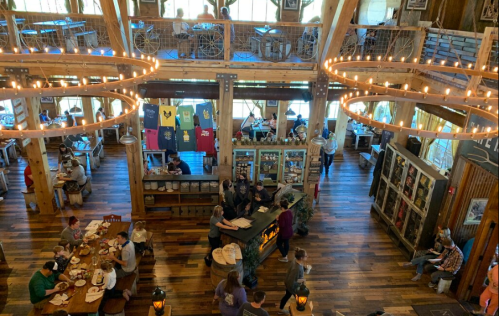 A bustling restaurant interior with wooden decor, people dining, and merchandise displayed on walls. Warm lighting enhances the atmosphere.