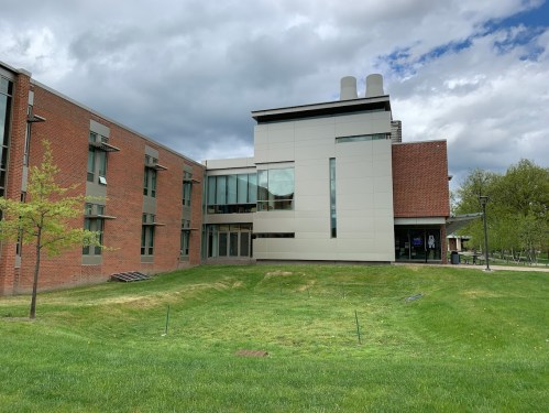 A modern building with a mix of brick and white exterior, surrounded by green grass and cloudy skies.
