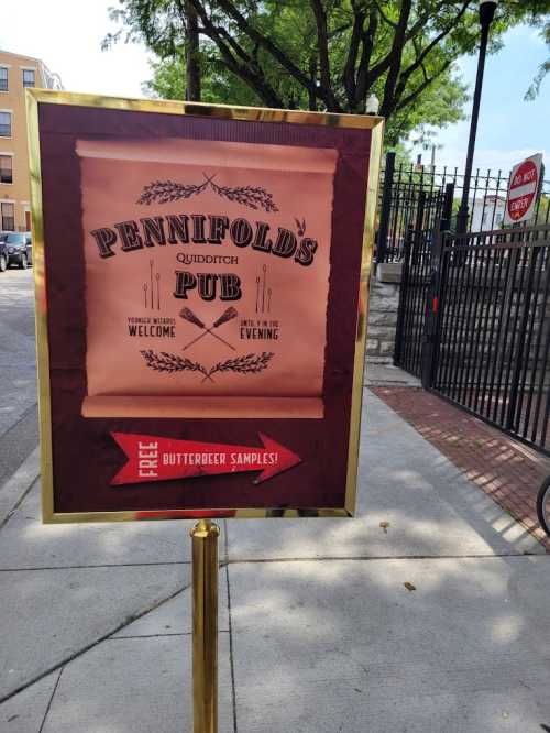 Sign for Pennifolds Pub offering free Butterbeer samples, with a Quidditch theme, set outside in a park-like area.