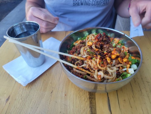 A bowl of spicy noodles topped with vegetables and meat, with chopsticks resting on the side, and a metal cup nearby.