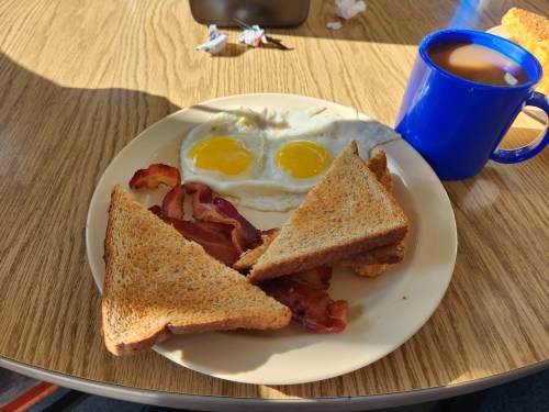A plate with two sunny-side-up eggs, crispy bacon, and toasted bread, alongside a blue mug of coffee.