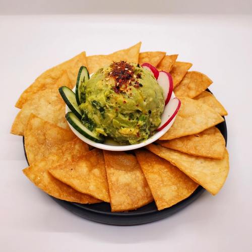 A bowl of guacamole topped with spices, surrounded by crispy tortilla chips and garnished with cucumber and radish slices.