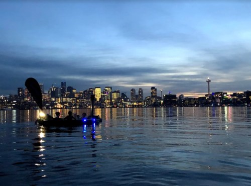A calm evening scene of a city skyline reflecting on water, with a small boat illuminated in the foreground.