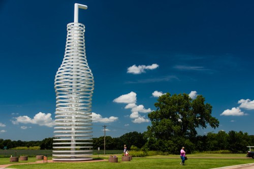 A large, spiraled sculpture resembling a bottle stands against a blue sky with clouds, surrounded by green grass and trees.