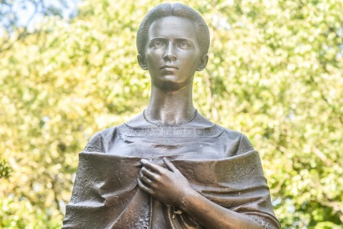 A bronze statue of a woman with a serious expression, holding her garment, set against a blurred green background.