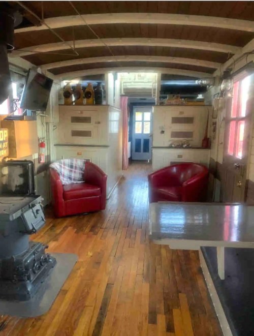 Cozy interior of a cabin with red chairs, wooden floors, and a stove, featuring a door leading outside.
