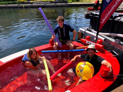 Three people play in a hot tub on a dock, using pool noodles and a beach ball, with water and greenery in the background.
