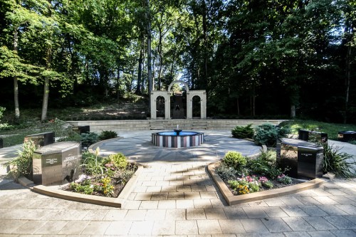 A serene memorial garden featuring a central fountain, surrounded by landscaped areas and stone monuments.