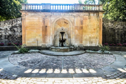 A stone fountain surrounded by a circular stone path and flower beds, set against a textured wall in a serene garden.