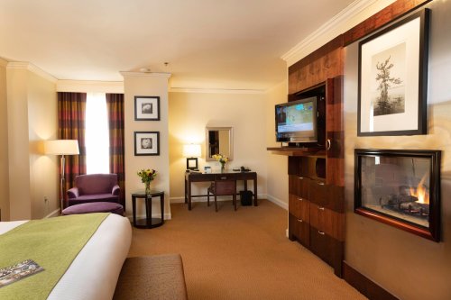 Cozy hotel room featuring a bed, armchair, desk, TV, and a fireplace, with warm lighting and modern decor.