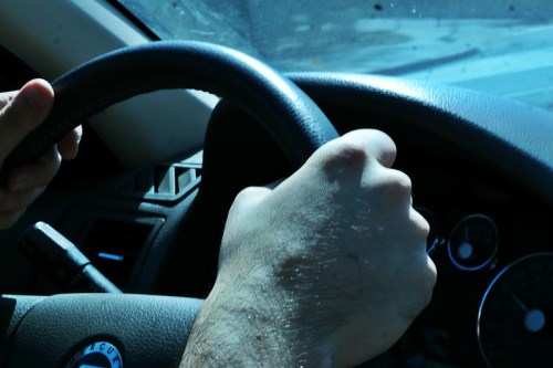 A close-up of hands gripping a steering wheel inside a car. Sunlight casts shadows on the dashboard.