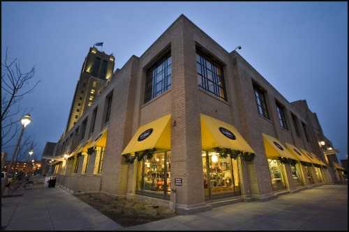 A corner building with yellow awnings at dusk, featuring large windows and a tall clock tower in the background.