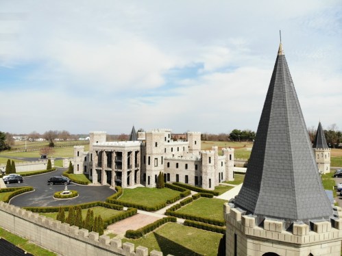 Aerial view of a large stone castle with turrets, surrounded by manicured lawns and a driveway.