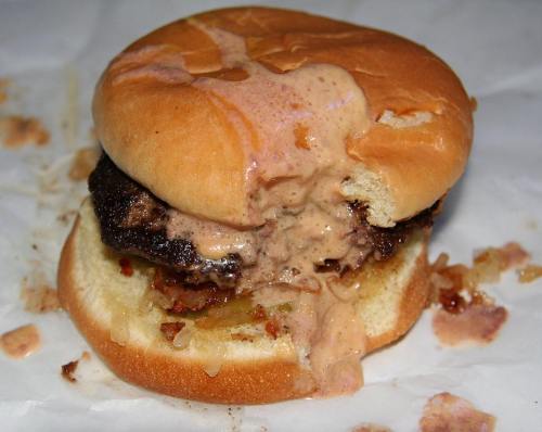 A messy hamburger with a juicy beef patty, melted cheese, and sauce dripping from a soft bun on a white wrapper.